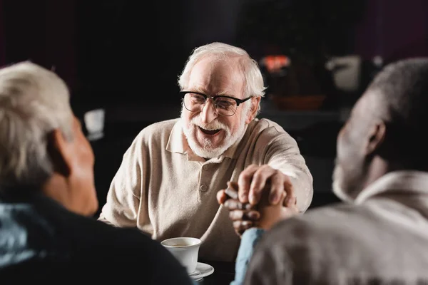 Excited senior man in eyeglasses joining hands with blurred multiethnic friends in bar — Stock Photo