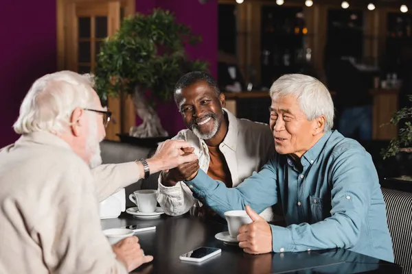 Feliz sénior multiétnicos amigos uniendo las manos mientras se sienta en la cafetería - foto de stock