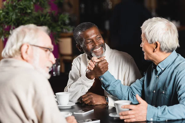 Cheerful multiethnic men joining hands near blurred senior friend in cafe — Stock Photo