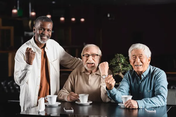 Amigos multiculturales senior mirando a la cámara y mostrando el gesto de ganar en la cafetería - foto de stock