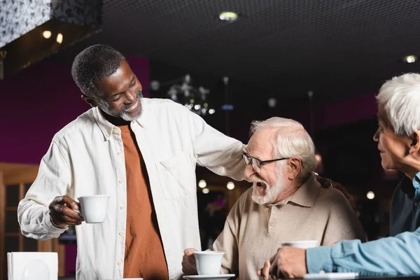 Sorrindo afro-americano homem com xícara de café de pé perto de amigos inter-raciais sênior rindo no café — Fotografia de Stock