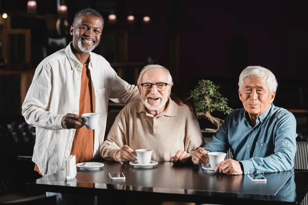 Amigos multiétnicos ancianos sonriendo a la cámara mientras beben café en el restaurante - foto de stock