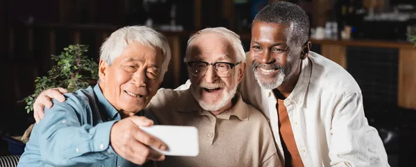 Lächelnder asiatischer Mann macht Selfie auf Smartphone mit älteren multiethnischen Freunden, Banner — Stockfoto