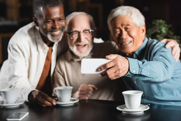 Joyful multiethnic friends taking selfie on mobile phone in cafe, blurred background — Stock Photo