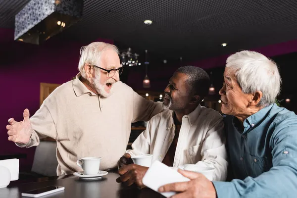Excited senior man showing wow gesture near happy multiethnic friends in cafe — Stock Photo
