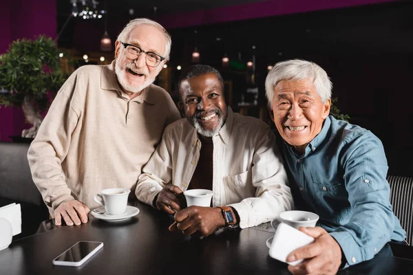 Senior and cheerful multicultural friends looking at camera during meeting in restaurant — Stock Photo