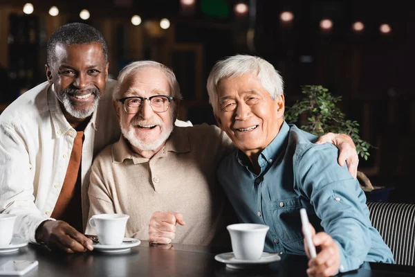 Amis multiculturels aînés embrasser et sourire à la caméra près des tasses à café dans le restaurant — Photo de stock