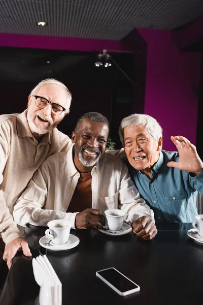 Счастливый азиатский мужчина машет рукой рядом со старшими многонациональными друзьями в кафе — стоковое фото