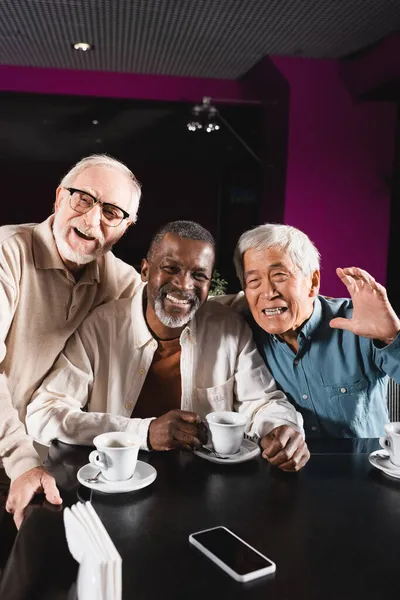 Feliz asiático homem acenando mão perto sênior multiétnico amigos no café — Fotografia de Stock