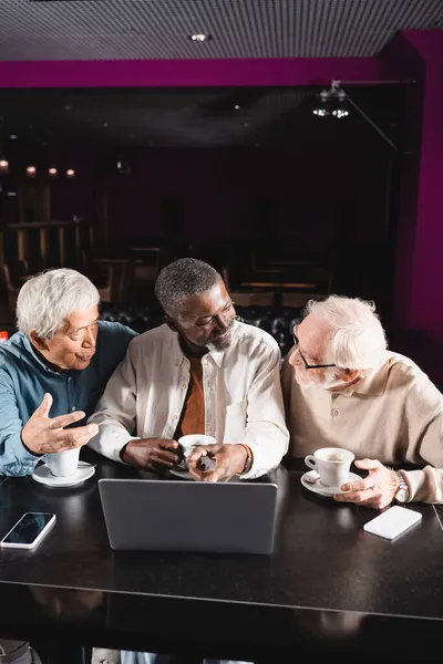 Hombre afroamericano mayor señalando a la computadora portátil durante la conversación con amigos multiétnicos en la cafetería — Stock Photo