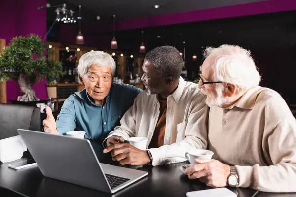 Mayores amigos interracial apuntando a la computadora portátil mientras habla en la cafetería - foto de stock