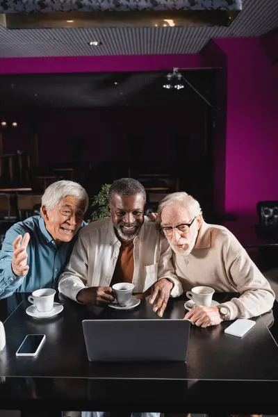 Joyful senior multicultural friends gesturing near laptop during video call in cafe — Stock Photo