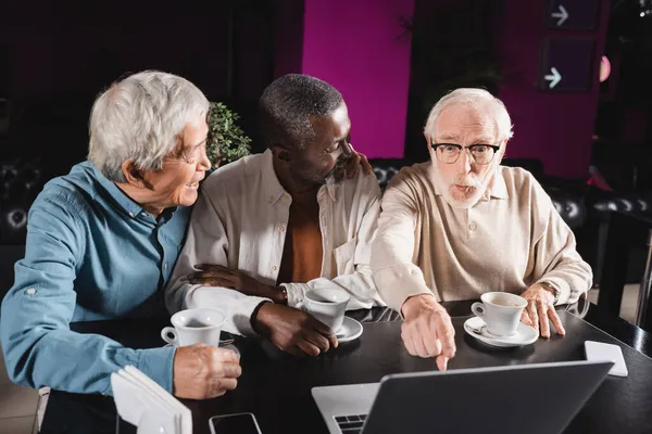 Homme âgé étonné pointant vers ordinateur portable près souriant amis multiethniques dans le café — Photo de stock