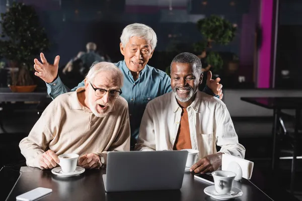 Senior asiático hombre gesto cerca multiétnico amigos durante vídeo llamada en portátil - foto de stock