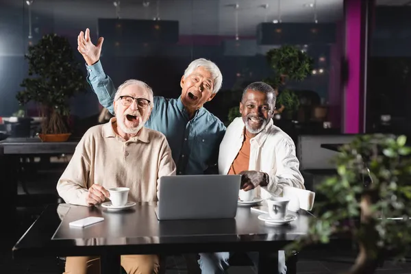 Alegre sénior amigos multiétnicos mirando la cámara cerca de la computadora portátil en la cafetería - foto de stock