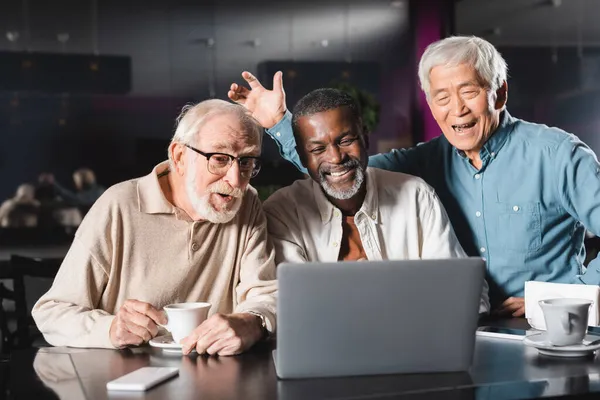 Alegre asiático hombre saludando mano cerca sénior multiétnico amigos durante vídeo llamada en portátil en café - foto de stock