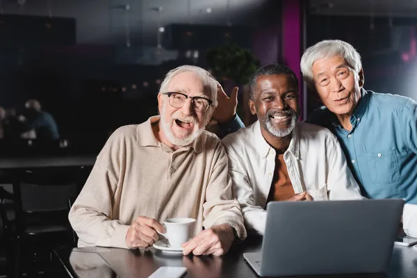 Amici multietnici anziani gioiosi che sorridono a macchina fotografica vicino a computer portatile in caffè — Foto stock