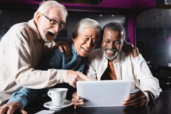 Asombrado hombre mayor apuntando a la tableta digital cerca de alegres amigos multiétnicos en la cafetería - foto de stock