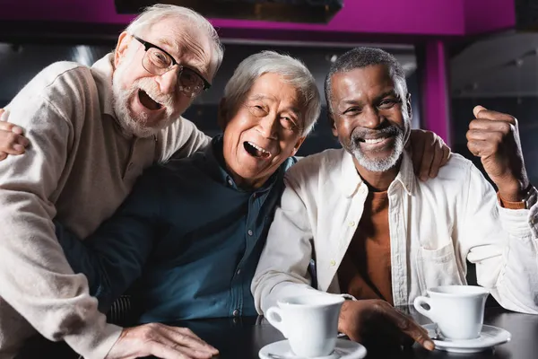 Smiling african american man showing rejoice gesture near laughing senior friends in cafe — Stock Photo