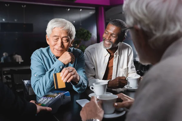 Senior asian man paying near credit card reader in cafe near multiethnic friends — Stock Photo