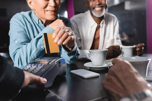 Vista recortada de hombre mayor con tarjeta de crédito cerca de la terminal de pago y hombre afroamericano en la cafetería - foto de stock