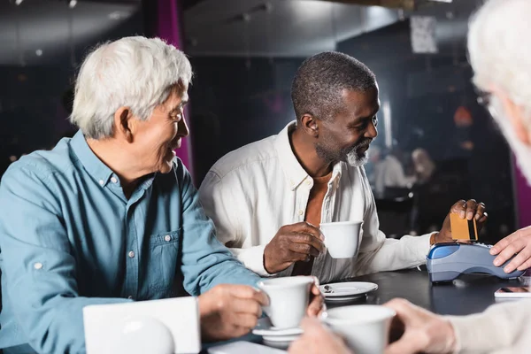 Afroamericano hombre pagar a través de lector de tarjetas de crédito cerca borrosa amigos en la cafetería - foto de stock