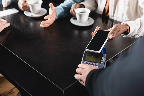 Vista cortada do homem americano africano pagando através do terminal com telefone celular perto de amigos turvos — Fotografia de Stock