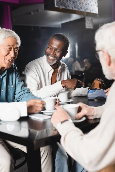 Glücklich afrikanisch-amerikanischer Mann zeigt auf Zahlungsterminal in der Nähe verschwommener multiethnischer Freunde — Stockfoto