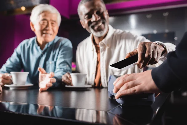 Blurred senior man paying through terminal with mobile phone near asian friend — Stock Photo