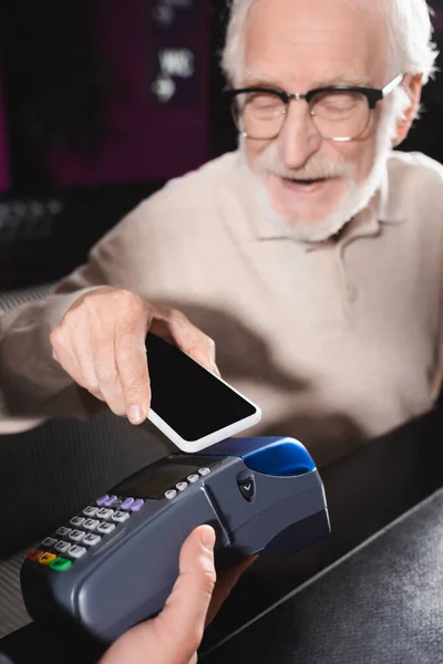 Hombre mayor en gafas de vista usando smartphone con pantalla en blanco mientras paga a través de terminal - foto de stock