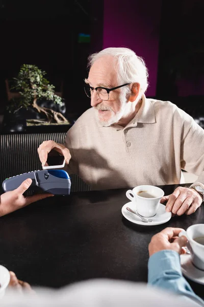 Hombre mayor en gafas que pagan con el teléfono celular a través de la terminal en el bar - foto de stock