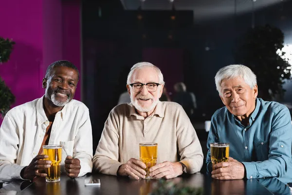 Feliz sénior amigos multiétnicos mirando a la cámara mientras está sentado en el pub con vasos de cerveza - foto de stock
