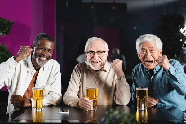 Feliz sénior amigos multiétnicos mostrando gesto de victoria mientras ve el campeonato de fútbol en el pub - foto de stock