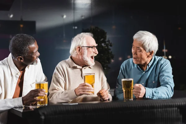 Hombre mayor riendo mientras bebe cerveza con amigos multiétnicos - foto de stock