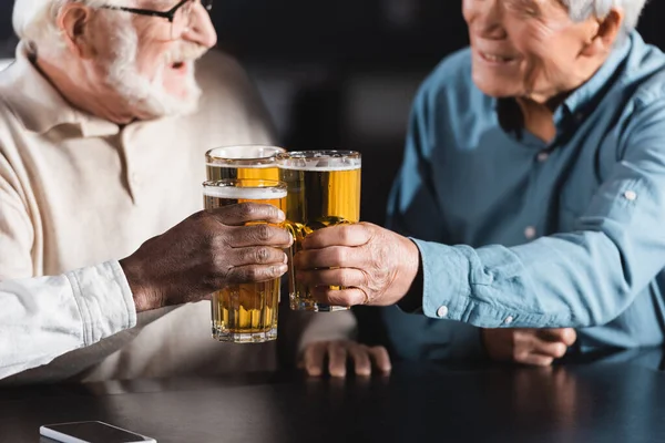 Amigos multiétnicos mayores tintineando vasos de cerveza mientras pasan tiempo en el pub en un fondo borroso - foto de stock