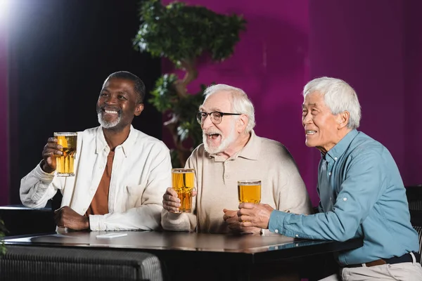 Веселые пожилые многонациональные друзья с бокалами пива смотрят футбольный матч в пабе — стоковое фото