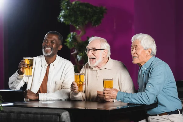 Joyful elderly multiethnic friends with glasses of beer watching football match in pub — Stock Photo