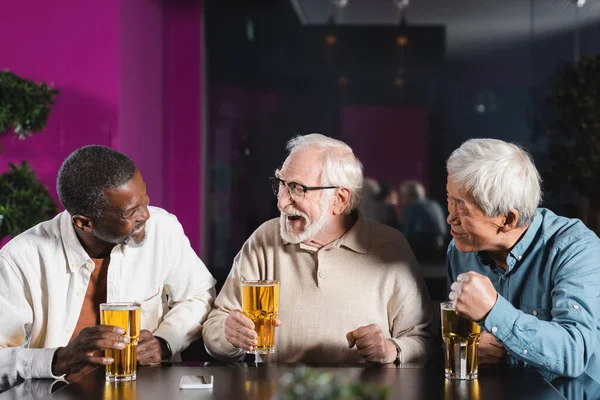 Amigos multiculturales ancianos emocionados hablando en el pub cerca de vasos de cerveza - foto de stock