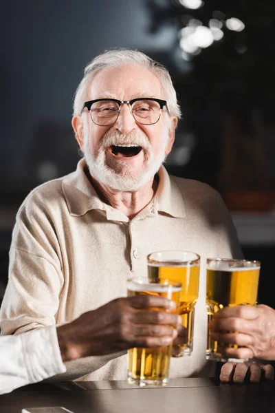 Senior man in eyeglasses laughing while clinking beer glasses with blurred interracial friends — Stock Photo