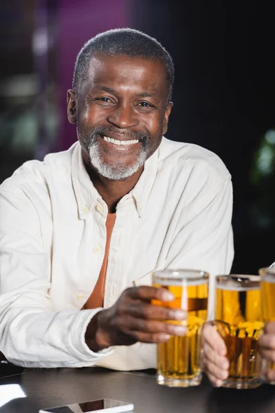 Älterer afrikanisch-amerikanischer Mann lächelt in die Kamera, während er mit Freunden verschwommene Biergläser klimpert — Stockfoto