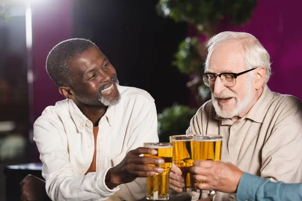 Feliz sénior amigos multiétnicos tintineo vasos de cerveza mientras pasar tiempo en el pub - foto de stock