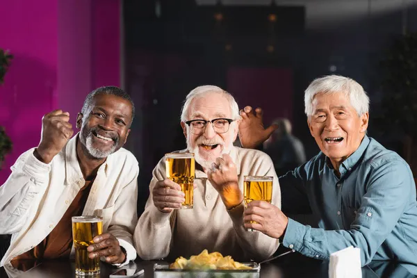 Joyeux amis multiculturels seniors avec des verres de bière montrant geste de victoire tout en regardant le match de football dans le pub — Photo de stock