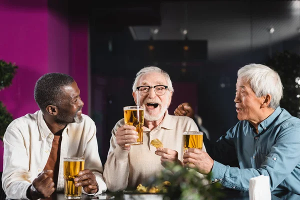 Веселый мужчина держит стакан пива рядом со старшими многонациональными друзьями в пабе — стоковое фото