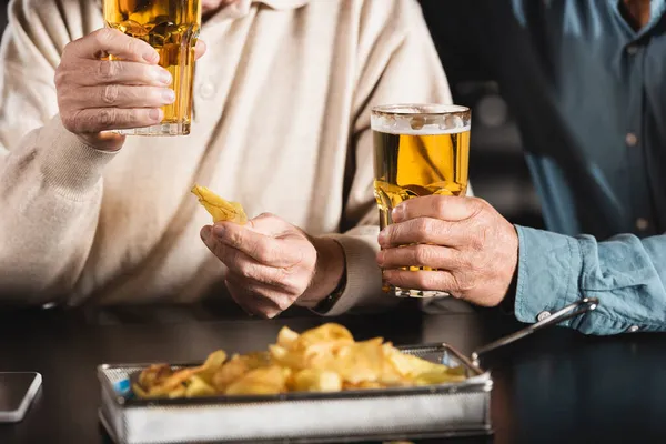Vista cortada de amigos seniores segurando copos de cerveja perto de chips borrados — Fotografia de Stock