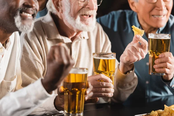 Cropped view of senior multiethnic friends eating chips and drinking beer in pub — Stock Photo