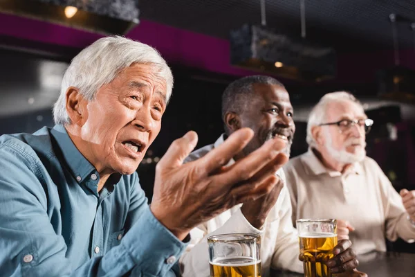 Обескураженный азиатский мужчина, указывающий рукой во время просмотра футбольного матча с улыбающимися межрасовыми друзьями в пабе — стоковое фото