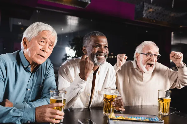 Excité homme criant et montrant geste de victoire tout en regardant le championnat de football avec des amis interracial senior — Photo de stock