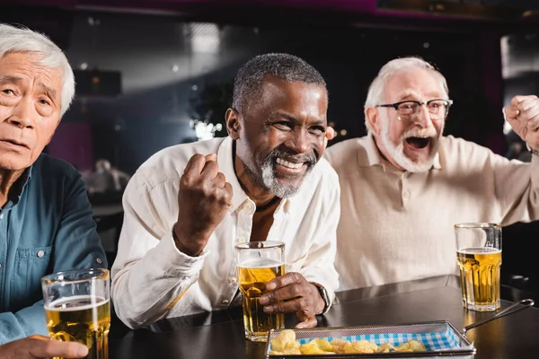 Emocionado interracial amigos mostrando ganar gesto mientras viendo fútbol cerca molesto asiático hombre - foto de stock