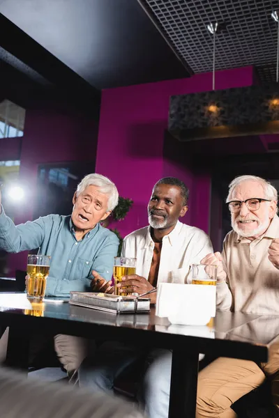 Tenso hombre mayor con los puños cerrados viendo el partido de fútbol con amigos multiétnicos en el pub - foto de stock