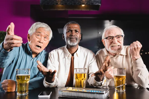 Emotivo anziani amici multiculturali guardando partita di calcio vicino a bicchieri di birra e patatine fritte in pub — Foto stock